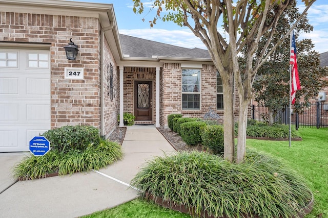 doorway to property featuring brick siding, a yard, roof with shingles, an attached garage, and fence