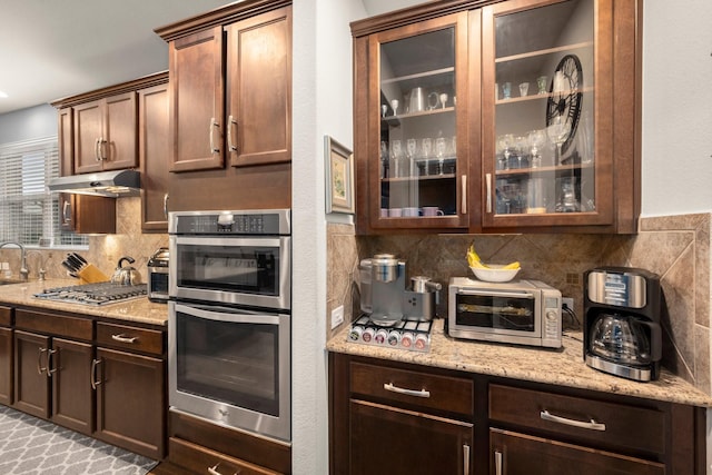kitchen featuring a toaster, under cabinet range hood, appliances with stainless steel finishes, backsplash, and glass insert cabinets