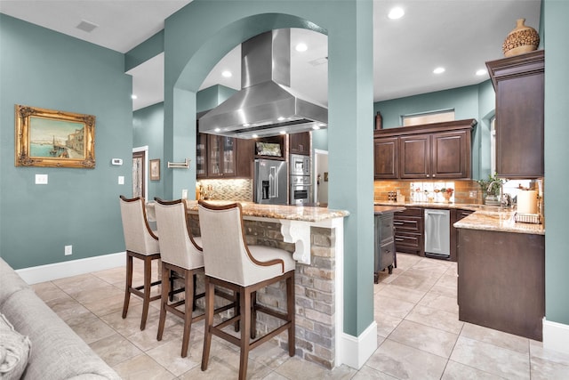 kitchen featuring a kitchen island, island range hood, dark brown cabinetry, stainless steel appliances, and light stone countertops