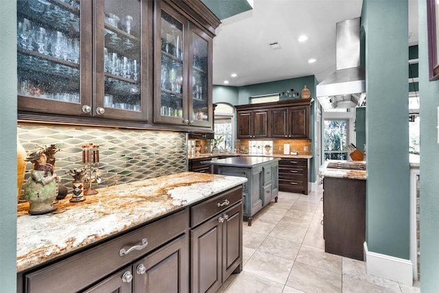 kitchen featuring dark brown cabinetry, extractor fan, light tile patterned floors, light stone countertops, and decorative backsplash