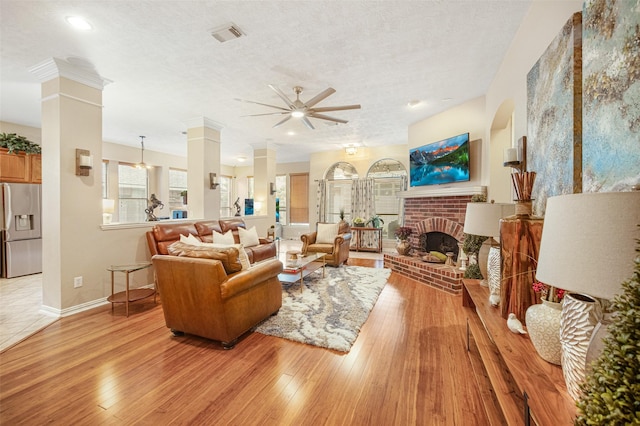 living room with a brick fireplace, light wood-type flooring, a textured ceiling, and ornate columns