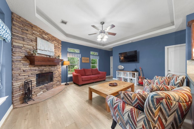 living room with a stone fireplace, light hardwood / wood-style flooring, ceiling fan, and a tray ceiling