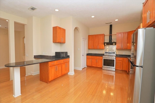 kitchen featuring wall chimney exhaust hood, a breakfast bar, appliances with stainless steel finishes, kitchen peninsula, and light hardwood / wood-style floors