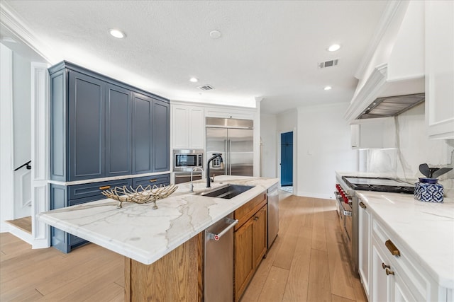 kitchen with sink, light stone counters, built in appliances, an island with sink, and white cabinets