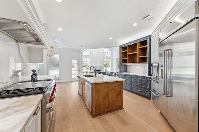 kitchen with sink, built in appliances, tasteful backsplash, light stone countertops, and an island with sink