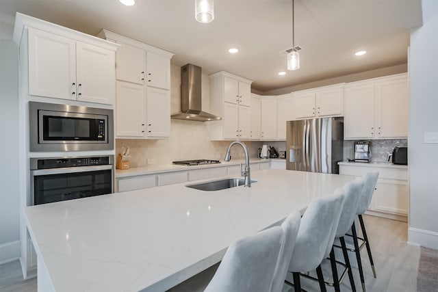 kitchen with wall chimney range hood, sink, stainless steel appliances, an island with sink, and decorative light fixtures