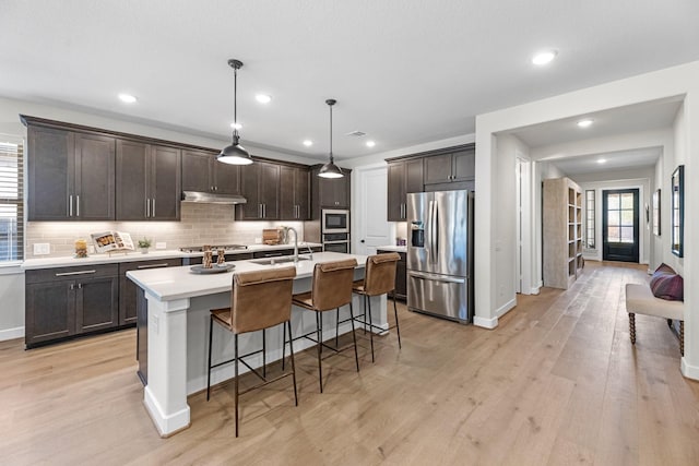 kitchen featuring decorative light fixtures, light hardwood / wood-style floors, dark brown cabinetry, stainless steel appliances, and a center island with sink