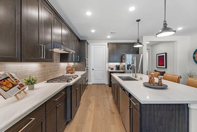 kitchen with sink, hanging light fixtures, light wood-type flooring, appliances with stainless steel finishes, and a kitchen island with sink