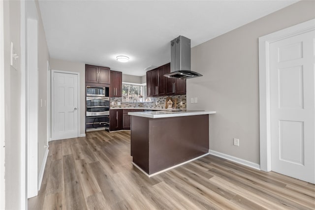 kitchen with appliances with stainless steel finishes, tasteful backsplash, island exhaust hood, kitchen peninsula, and light hardwood / wood-style flooring