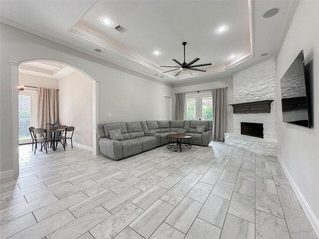 living room with a raised ceiling, ornamental molding, a stone fireplace, and ceiling fan