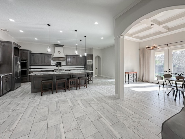 kitchen featuring dark brown cabinetry, coffered ceiling, custom range hood, and an island with sink