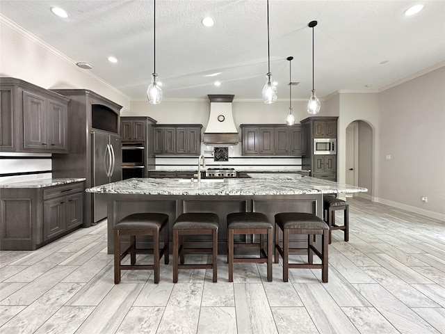 kitchen featuring arched walkways, a large island, dark brown cabinetry, built in appliances, and premium range hood