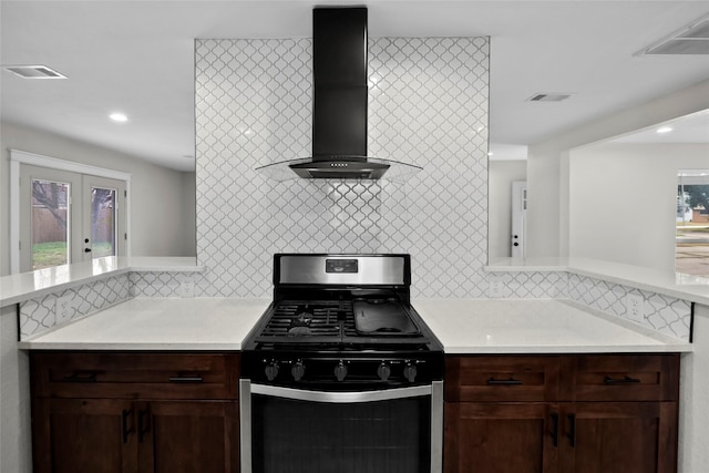 kitchen featuring stainless steel gas stove, dark brown cabinetry, decorative backsplash, kitchen peninsula, and wall chimney exhaust hood