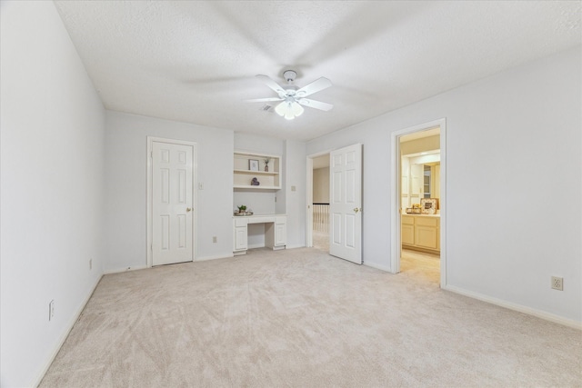 unfurnished bedroom featuring ensuite bathroom, built in desk, ceiling fan, light carpet, and a textured ceiling