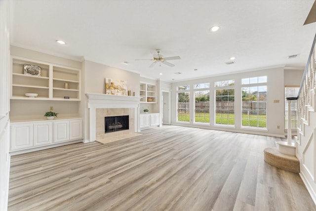 unfurnished living room featuring built in features, a tile fireplace, ceiling fan, ornamental molding, and light wood-type flooring