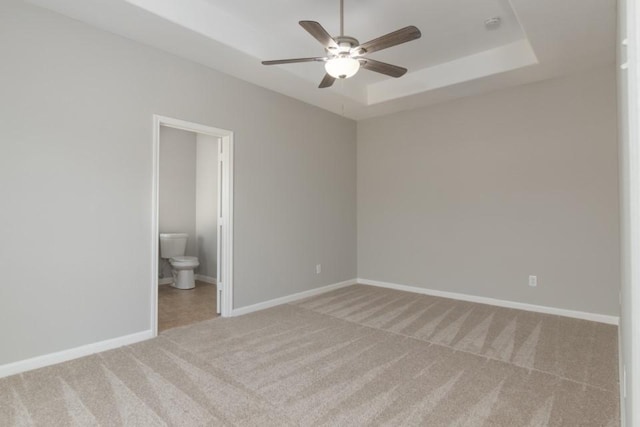 unfurnished bedroom with ceiling fan, ensuite bath, a tray ceiling, and carpet