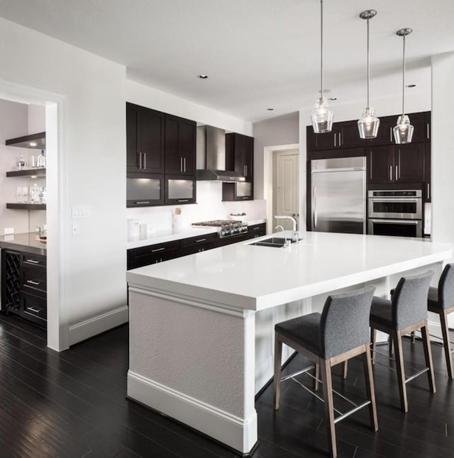 kitchen featuring wall chimney range hood, sink, appliances with stainless steel finishes, hanging light fixtures, and an island with sink