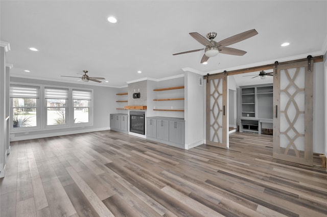 unfurnished living room featuring light hardwood / wood-style flooring, ornamental molding, ceiling fan, a barn door, and built in shelves