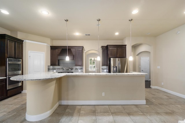 kitchen with wall chimney exhaust hood, dark brown cabinetry, hanging light fixtures, appliances with stainless steel finishes, and a large island