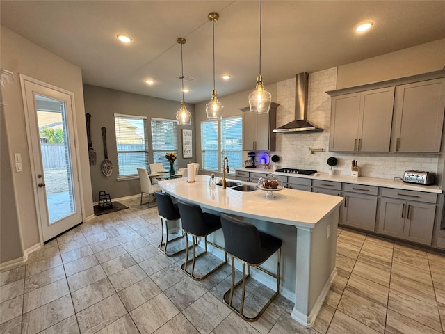 kitchen featuring a breakfast bar, hanging light fixtures, black gas cooktop, a center island with sink, and wall chimney exhaust hood