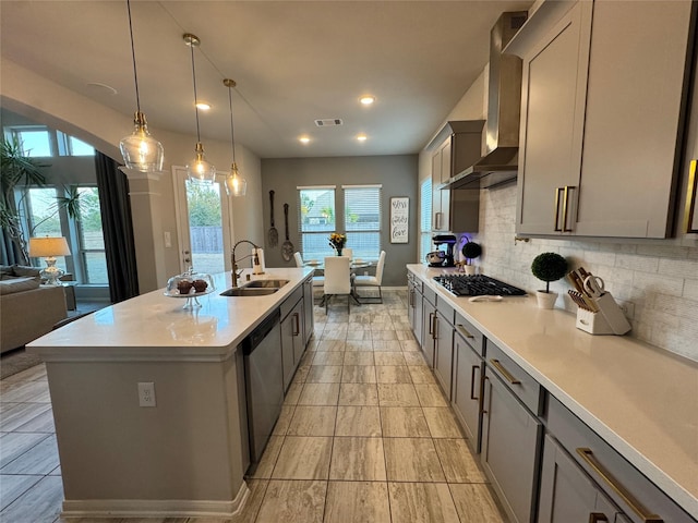 kitchen with wall chimney exhaust hood, sink, hanging light fixtures, appliances with stainless steel finishes, and an island with sink