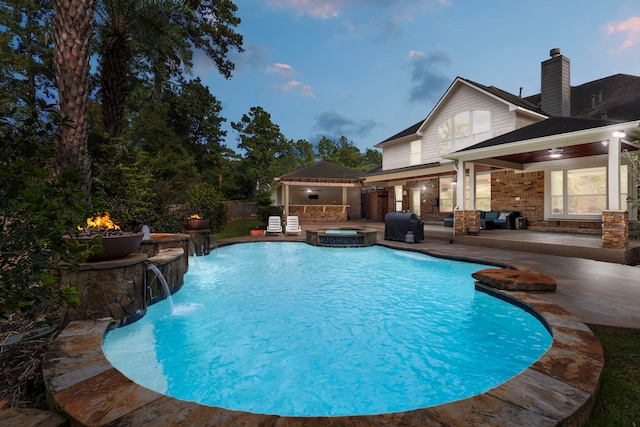 pool at dusk featuring an in ground hot tub, pool water feature, a patio area, and an outdoor fire pit