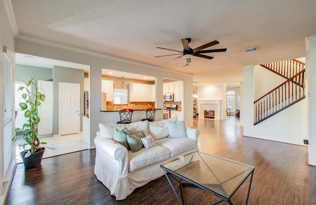 living room with dark wood-type flooring, a tile fireplace, ceiling fan, ornamental molding, and a textured ceiling