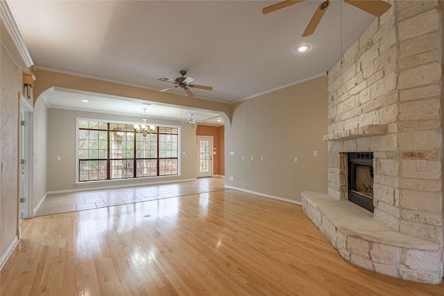 unfurnished living room with crown molding, light wood-style floors, a stone fireplace, baseboards, and ceiling fan with notable chandelier