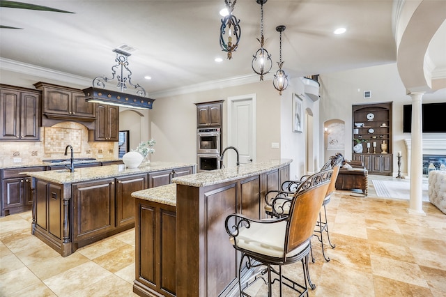 kitchen with dark brown cabinets, hanging light fixtures, a center island with sink, and ornate columns