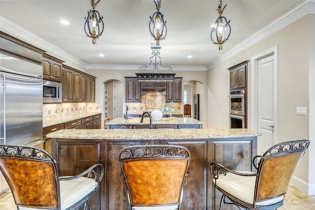 kitchen featuring decorative light fixtures, a kitchen island with sink, built in appliances, dark brown cabinetry, and light stone countertops