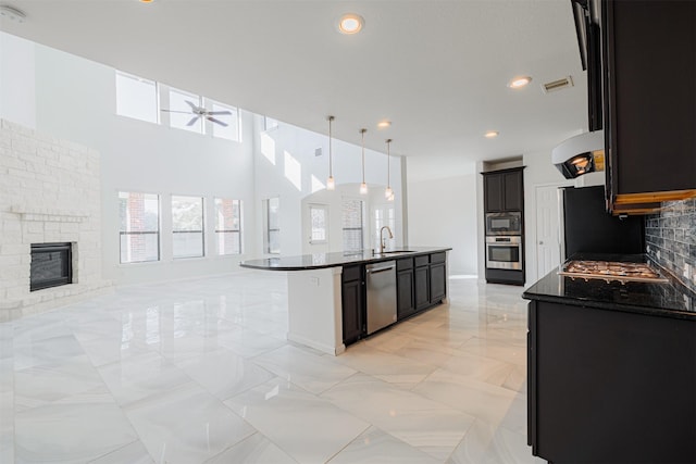 kitchen with a stone fireplace, sink, hanging light fixtures, a center island with sink, and stainless steel appliances