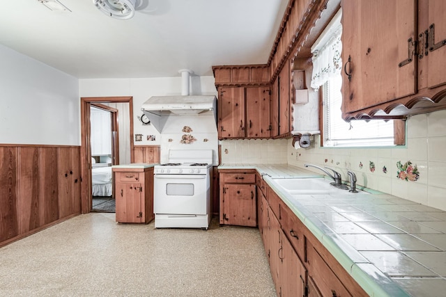 kitchen with sink, white gas range oven, ventilation hood, tile countertops, and wood walls