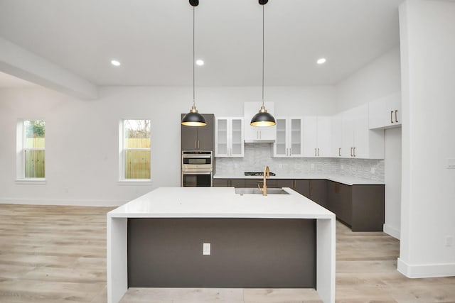 kitchen with white cabinets, glass insert cabinets, decorative light fixtures, a kitchen island with sink, and light countertops