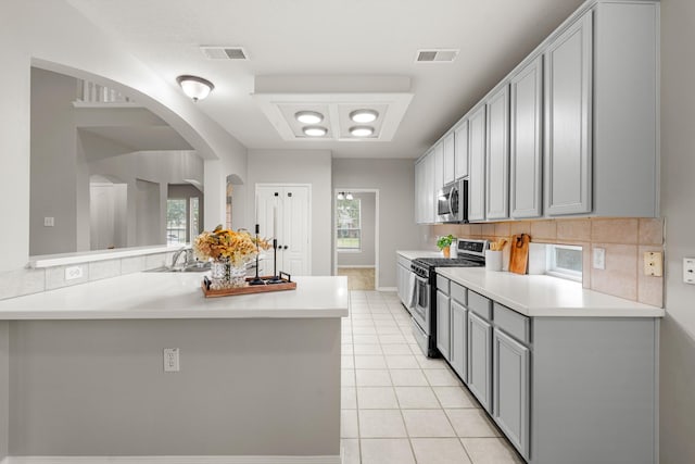 kitchen with light tile patterned flooring, gray cabinets, kitchen peninsula, stainless steel appliances, and backsplash