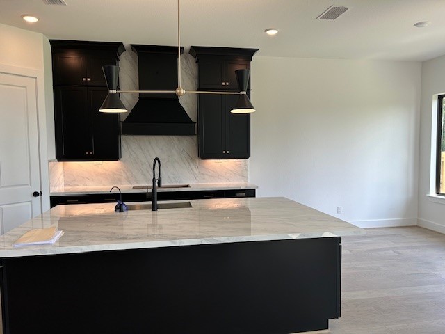 kitchen with sink, a kitchen island with sink, backsplash, light stone countertops, and custom exhaust hood