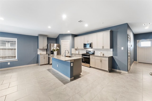 kitchen featuring sink, appliances with stainless steel finishes, light stone countertops, an island with sink, and decorative backsplash