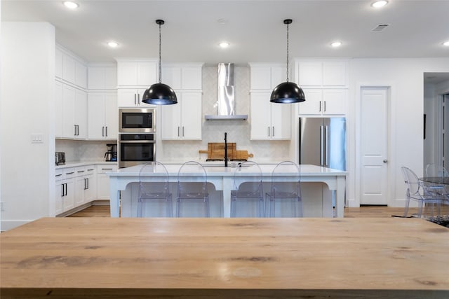 kitchen featuring pendant lighting, appliances with stainless steel finishes, a kitchen breakfast bar, a center island with sink, and wall chimney exhaust hood