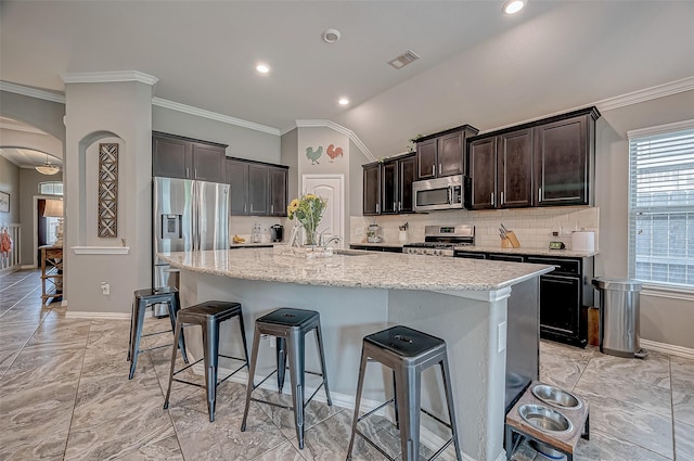 kitchen with a kitchen bar, light stone counters, ornamental molding, appliances with stainless steel finishes, and an island with sink