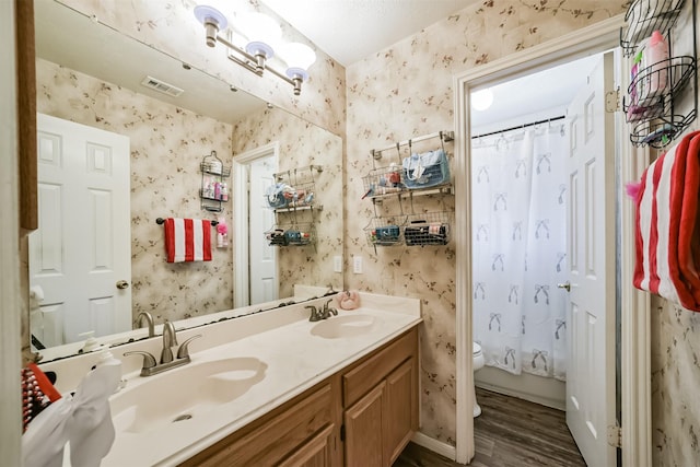 bathroom with toilet, wallpapered walls, double vanity, and a sink