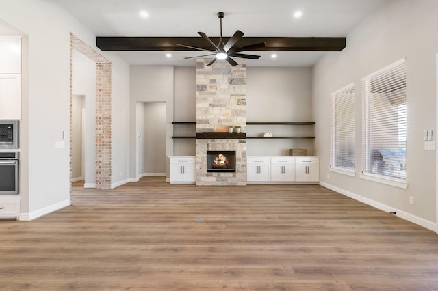 unfurnished living room with beamed ceiling, ceiling fan, a stone fireplace, and light hardwood / wood-style flooring