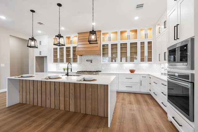 kitchen featuring custom exhaust hood, stainless steel microwave, a kitchen island with sink, light hardwood / wood-style floors, and white cabinets