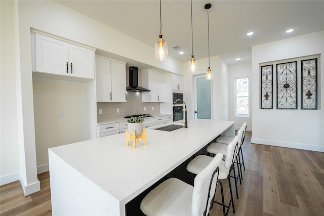 kitchen featuring white cabinetry, wall chimney range hood, hanging light fixtures, and a center island with sink