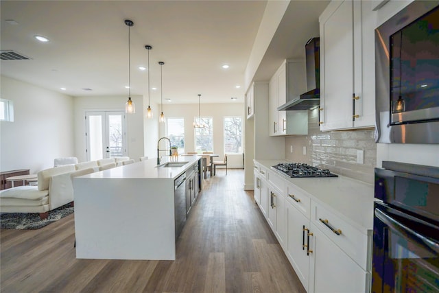 kitchen featuring white cabinetry, hanging light fixtures, stainless steel appliances, a kitchen island with sink, and wall chimney range hood