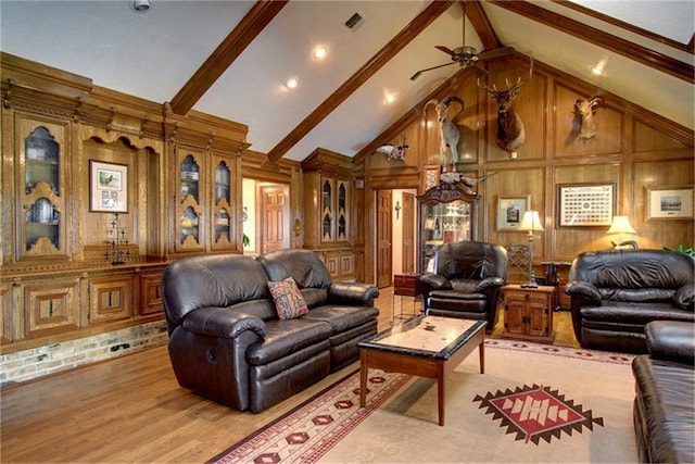 living room with ceiling fan, beam ceiling, high vaulted ceiling, light wood-type flooring, and wood walls