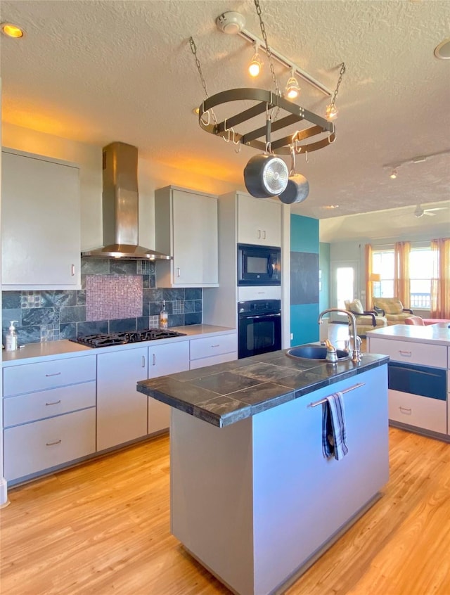 kitchen featuring wall chimney range hood, light wood-type flooring, sink, and black appliances