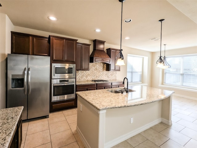 kitchen featuring an island with sink, appliances with stainless steel finishes, light stone counters, premium range hood, and a sink