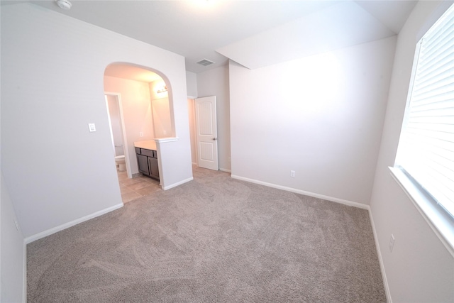 unfurnished bedroom with arched walkways, light carpet, visible vents, baseboards, and ensuite bath