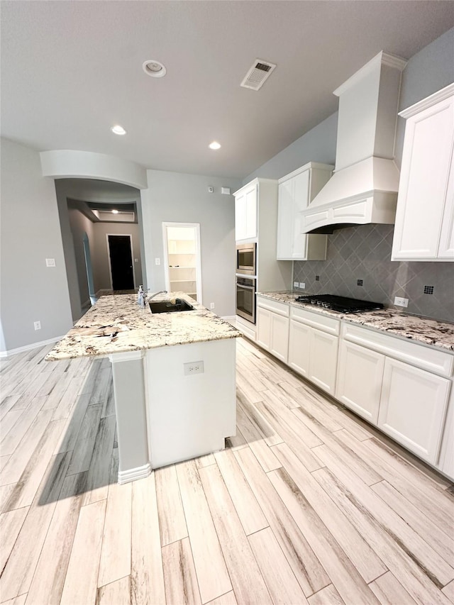 kitchen with custom exhaust hood, light stone counters, appliances with stainless steel finishes, an island with sink, and white cabinets