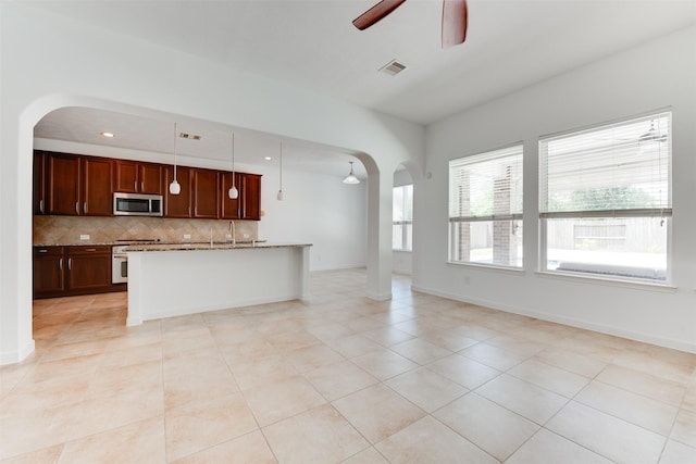 kitchen with ceiling fan, a kitchen island with sink, tasteful backsplash, light stone countertops, and decorative light fixtures