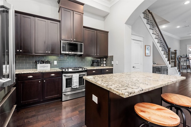 kitchen with dark brown cabinets, stainless steel appliances, a center island, light stone counters, and ornamental molding